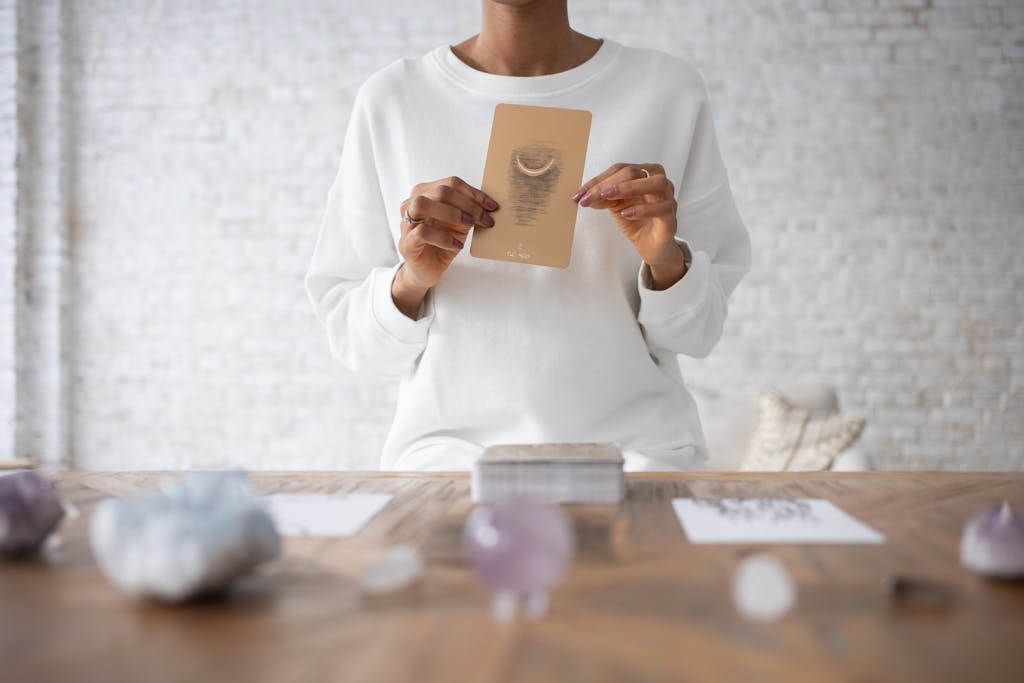 A person wearing white holds a tarot card with gemstones on a wooden table for divination.