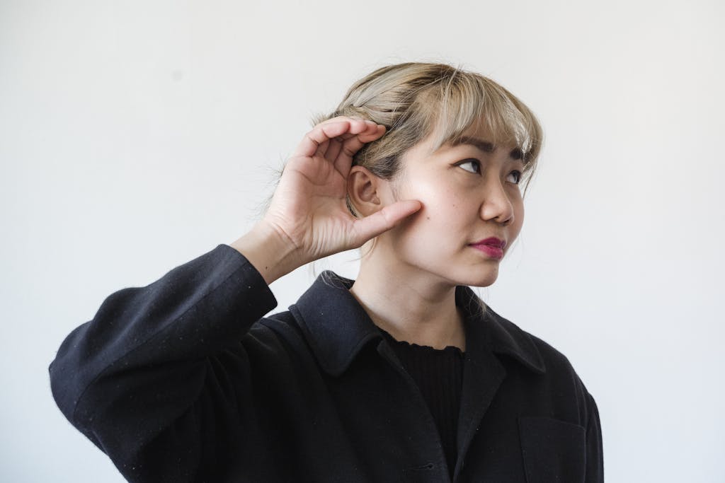 A thoughtful adult woman with blonde hair posing indoors, hand over ear.