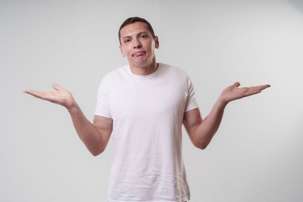 An adult man wearing a white t-shirt shrugs against a neutral background, conveying confusion or indifference.