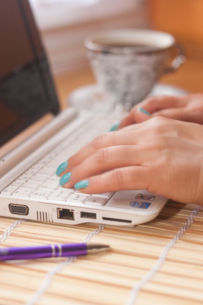 Close-up of a woman's hands typing on a white laptop with coffee beside.