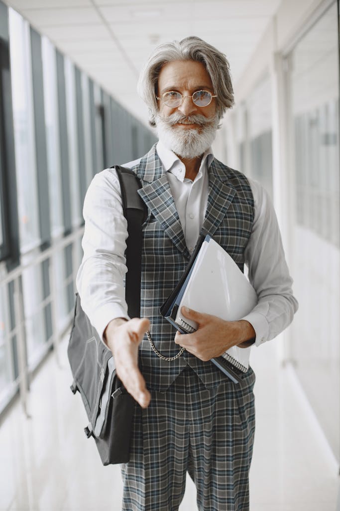 Mature bearded businessman with documents, smiling and extending hand for a handshake in a modern office hallway.