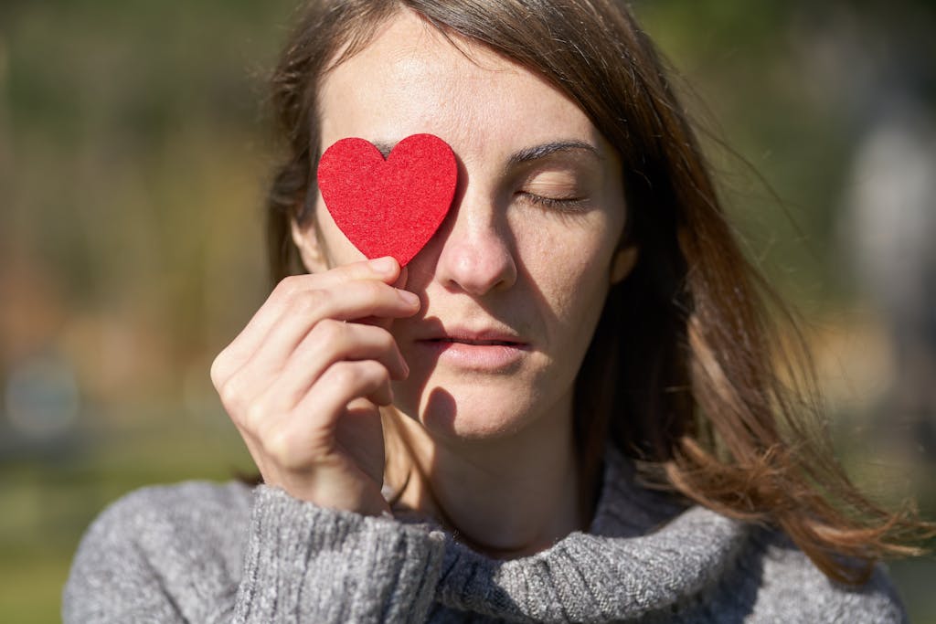 Woman holding a red heart symbol in front of her eye, enjoying a sunny outdoor day.