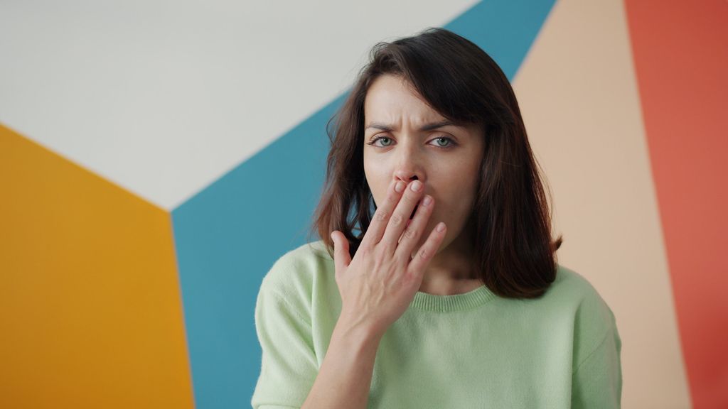 Woman covers her mouth against a colorful geometric wall, expressing surprise or yawn.